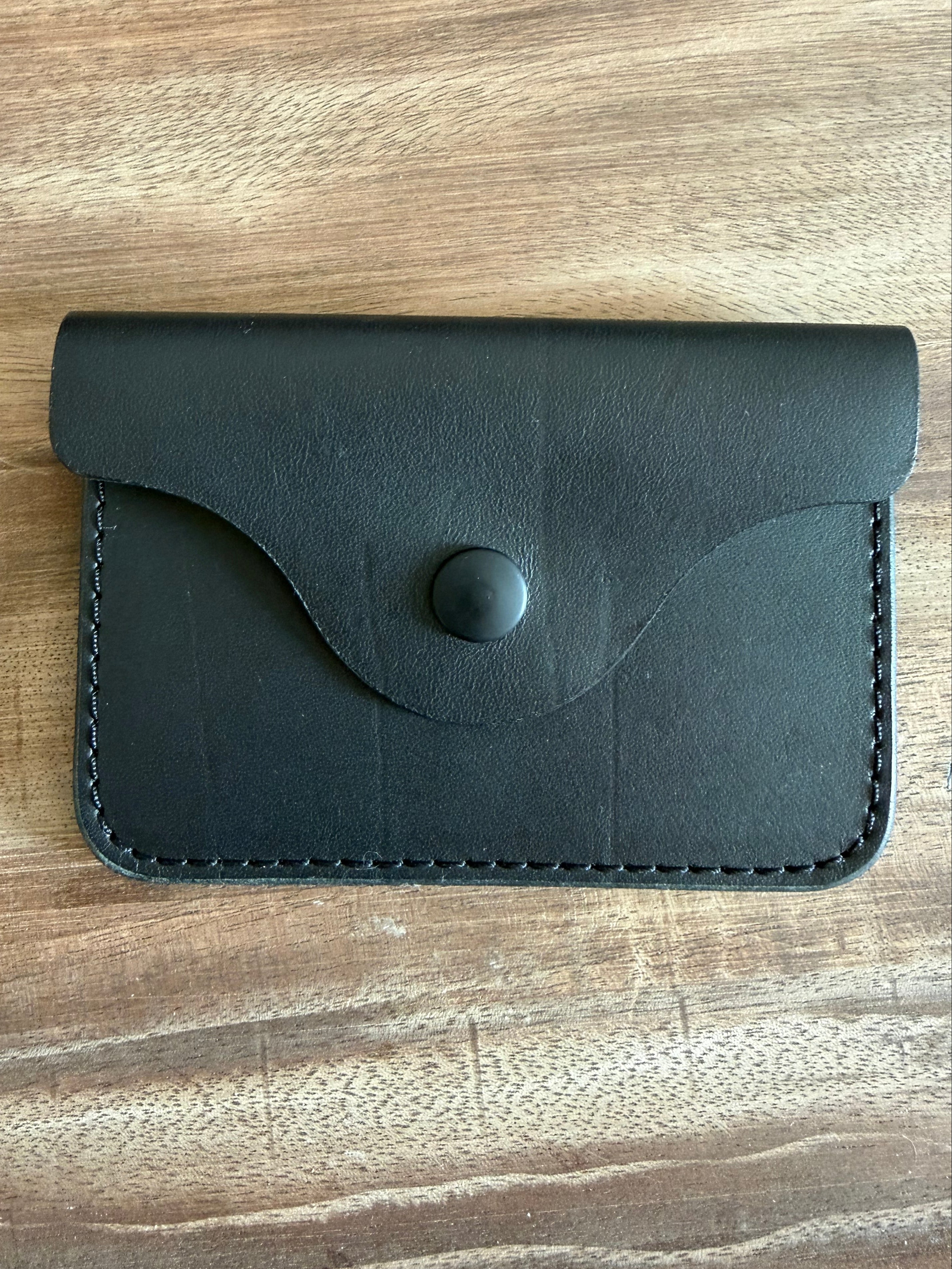 A small black leather card holder or wallet with a flap closure and snap button, photographed from above on a wooden surface.