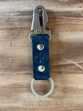 A dark blue leather keychain with silver-tone metal hardware, including a split ring, snap buttons, and a carabiner clip, displayed on a wooden surface.